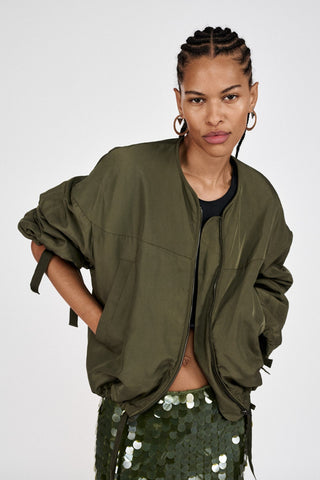 Bomberjacke in army green mit lässigen Bändern am Bund und Armabschluss. Das Model trägt sie offen über einem kurzen schwarzen Top und einem Pailleten Jupe.