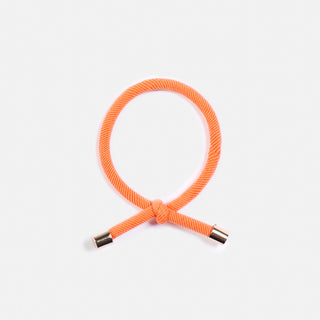 man sieht ein orangenes Haargummi der Schweizer Marke AYRE. Es hat einen Knopf im Haargummi und die Enden haben goldene Abschlüsse.