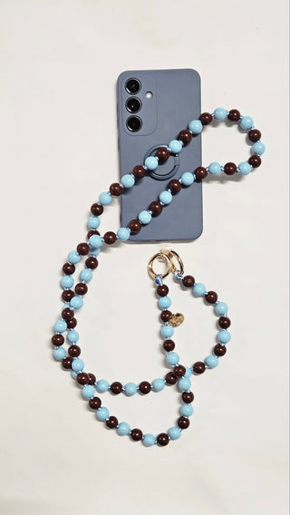 Man sieht die Handykette der Schweizer Marke AYRE. Eine Handykette aus Holzperlen welche von Hand zusammengeknüpft wurden und sie ist in der Farbkombination Hellblau und Brown. mit goldenen Karabinern um am Handpatch anzubringn.