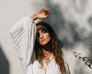 Man sieht ein Model in der Sonne mit einem schönen Broderie Anglaise Kleid in weiss, von der schweizer Marke AYRE. Lange weite Ärmel. 