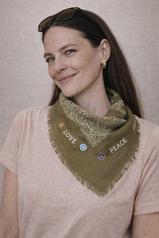 lachendes Model zeigt den Monsterli Schal der schweizer Marke AYRE.  Hier in der Farbe Olive. Der Schal hat im inneren des Quadrates ein klassisches Paisley Muster, aussen herum hat er süsse Stickereien die gute Laune machen.