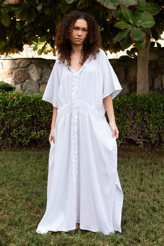 Fliessendes Maxikleid aus 100 % Baumwoll-Musselin in reinem Weiss. Weite Silhouette mit V-Ausschnitt, strukturierter Zierkante und vertikaler Textur. Leicht, atmungsaktiv und vielseitig – ideal für Sommer, Reisen und moderne Resortlooks. One Size.