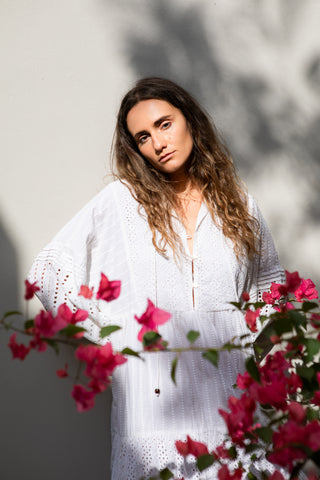 Luftiges Boho-Kleid aus 100 % Baumwolle mit feinen Stickereien und Stufenschnitt. Fliessende Silhouette, weite Ärmel und elegante Details – ideal für Sommer, Resortwear und entspannte Alltagsmomente. One Size, leicht und vielseitig kombinierbar. Wunderschönes Moodbild. Model hinter Blumen.